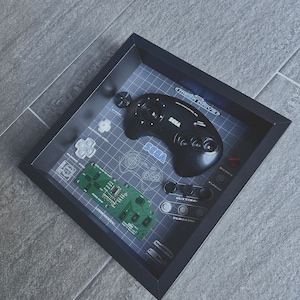 Könnte beinhalten: Ein schwarzer Sega Genesis-Controller und eine Platine, die in einem schwarzen Rahmen mit einem Gittermuster und dem Text "Sega Genesis" in der oberen rechten Ecke ausgestellt sind.