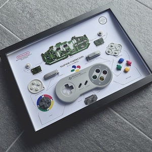 Könnte beinhalten: Eine gerahmte Darstellung von zerlegten Teilen eines Super Nintendo Entertainment System-Controllers. Die Teile umfassen die Platine, die Knöpfe und ein Gummipolster. Die Anzeige enthält auch den Text "SUPER FAMICOM" und "Nintendo SUPER FAMICOM".