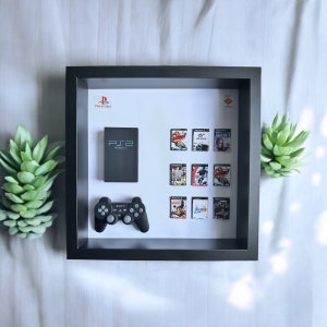 Op de afbeelding: Een zwarte, ingelijste schaduwdoos met een PlayStation 2-console, een zwarte controller en een verzameling PlayStation 2-spelcartridges. De doos heeft een witte achtergrond en de tekst "PlayStation" is in de linkerbovenhoek gedrukt.