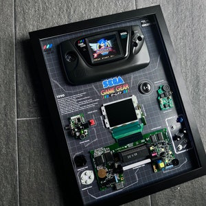 Op de afbeelding: Een ingelijst display van een gedemonteerde Sega Game Gear draagbare videogameconsole. De display bevat de hoofdprintplaat van de console, het scherm, de knoppen en andere componenten. De console wordt getoond met het spel Sonic the Hedgehog geladen.