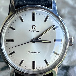 Pode incluir: Um relógio de pulso Omega Genève prateado com mostrador redondo, fundo metálico prateado, marcadores de hora pretos e ponteiros pretos. O relógio inclui uma pulseira de couro preta e exibe o logotipo da Omega e o texto T SWISS MADE T.