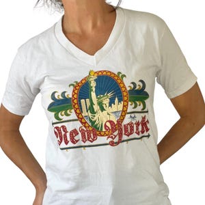 May include: White V-neck t-shirt featuring a colorful graphic of the Statue of Liberty and the text "New York". The graphic is set within a circular design with red, gold, blue, and green accents. The shirt is made of a soft material.