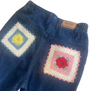 May include: Dark blue denim jeans with two crocheted patches on the back pockets. One patch features a yellow flower, and the other has a red rose. The jeans have a brown leather tag that says "GITANO".