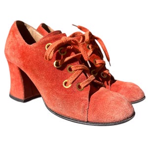 May include: A pair of orange suede lace-up heeled shoes. The shoes have a rounded toe, thick block heels, and gold-tone eyelets. The laces are the same color as the shoes. The shoes are set against a white background.