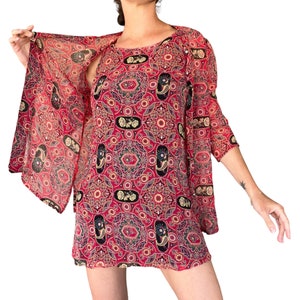 Pode incluir: Um vestido túnica vermelho com um padrão paisley em preto, dourado e verde. O vestido tem um decote redondo e mangas largas e fluidas. O tecido parece transparente, com uma queda leve.