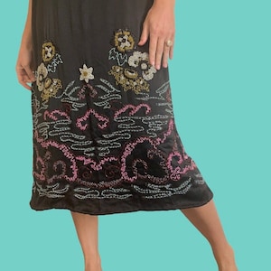 May include: A black dress with floral embroidery in gold, white, and pink. The dress features a knee-length hemline and is adorned with intricate beaded details. The design includes a combination of embroidered flowers and abstract patterns.