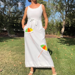 May include: White sleeveless maxi dress with a textured pattern. The dress features a tie waist, pockets, and two embroidered beach umbrellas in yellow, orange, and green. The hemline is trimmed with white pom-poms.