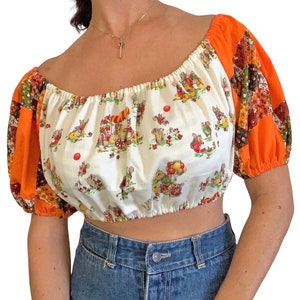May include: A cropped off-the-shoulder top with a whimsical print of cartoon animals and a patchwork design on the sleeves. The top features a white base with a repeating pattern of animals, red balloons, and a circus cart. The sleeves are orange and patterned.