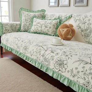 May include: A cream-colored sofa cover featuring a black floral line-art pattern and a light green ruffled trim. The set includes a square pillow and a rectangular pillow, both with matching floral prints and green ruffled edges.