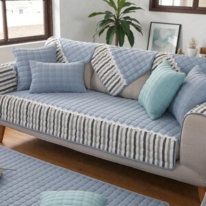 May include: A light blue quilted sofa cover and matching throw pillows. The cover features a striped border with white lace trim. Several throw pillows in varying shades of blue and a matching rug are also visible.