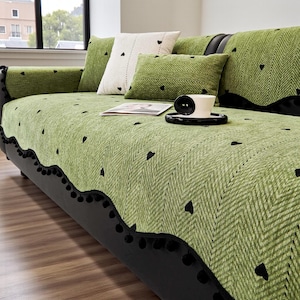 May include: A green sofa cover with a herringbone pattern and black heart accents. The cover has a scalloped edge with black pom-poms. Decorative pillows in green and white with heart designs are on the sofa. A cup and saucer sit on the cover.