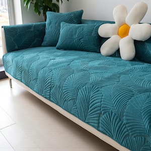 Puede incluir: Un sofá turquesa con cojines a juego y un gran cojín con forma de margarita. El sofá y los cojines tienen un diseño de hojas acolchadas. El sofá tiene un marco blanco y patas plateadas.