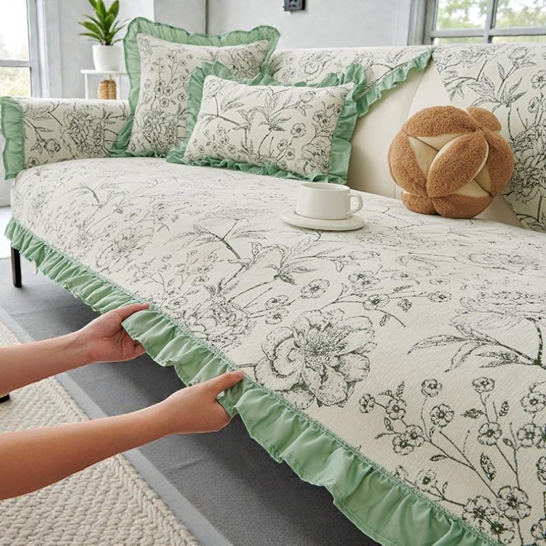 May include: A cream-colored sofa cover featuring a black floral line-art pattern and light green ruffled edges. The set includes matching square and rectangular throw pillows, along with a round, textured brown decorative cushion.