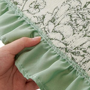 May include: A textile item featuring a cream-colored fabric with a dark green floral line-art pattern. The edge is finished with a gathered, light sage green fabric ruffle attached with a visible white serged stitch seam.