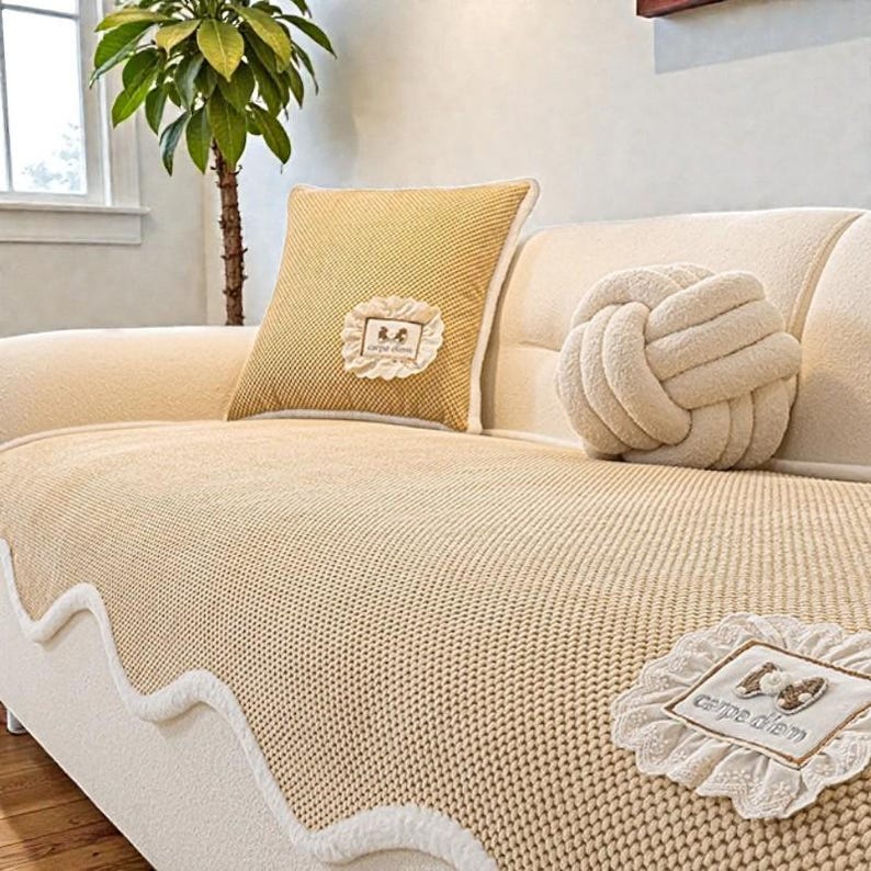 Pu&ograve; includere: Un divano beige con una coperta lavorata a maglia abbinata e cuscini decorativi. Un cuscino quadrato giallo con un'applicazione in pizzo bianco. Sono visibili anche un cuscino rotondo annodato e un'applicazione 'carpe diem'.