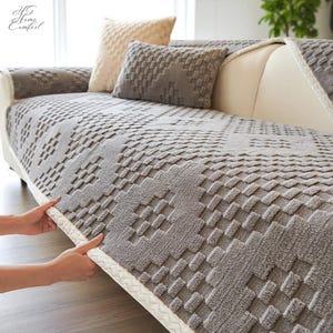 May include: A gray sofa cover with a textured geometric pattern, featuring matching throw pillows. The cover is being adjusted, showcasing its fit and design. The Home Comfort logo is visible in the upper left corner.