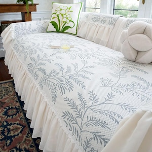 May include: A white sofa cover featuring a blue floral pattern and a ruffled skirt. The sofa has a matching cushion with a green floral design and a white decorative cushion. The sofa is in a living room setting.