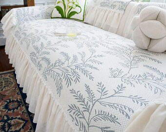 May include: A white sofa cover with a blue floral pattern and a ruffled skirt. The sofa cover has a matching pillow with a green floral design and a white decorative pillow. The sofa is in a living room setting.