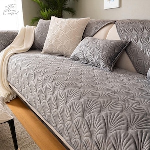 May include: A grey sofa cover featuring a fan-shaped design, with matching cushions. A cream-coloured blanket is draped over the arm. The sofa is set against a neutral backdrop, creating a cosy and inviting atmosphere.