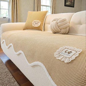 Pu&ograve; includere: Un divano beige con una fodera in maglia e cuscini decorativi. &Egrave; visibile un cuscino quadrato giallo con bordatura in pizzo bianco e un cuscino a forma di nodo. La fodera del divano ha un bordo bianco ondulato.