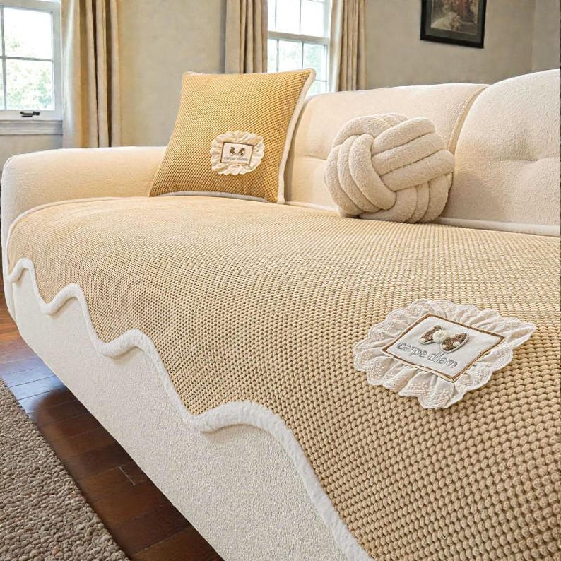 Pu&ograve; includere: Un divano beige con una fodera in maglia e cuscini decorativi. &Egrave; visibile un cuscino quadrato giallo con bordatura in pizzo bianco e un cuscino a forma di nodo. La fodera del divano ha un bordo bianco ondulato.