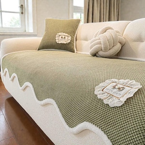 Pu&ograve; includere: Un divano color crema con un copriletto in maglia verde e un cuscino abbinato. Il copriletto ha un bordo bianco ondulato. Sono visibili anche un cuscino decorativo a nodo e una toppa quadrata con la scritta "carpe diem".