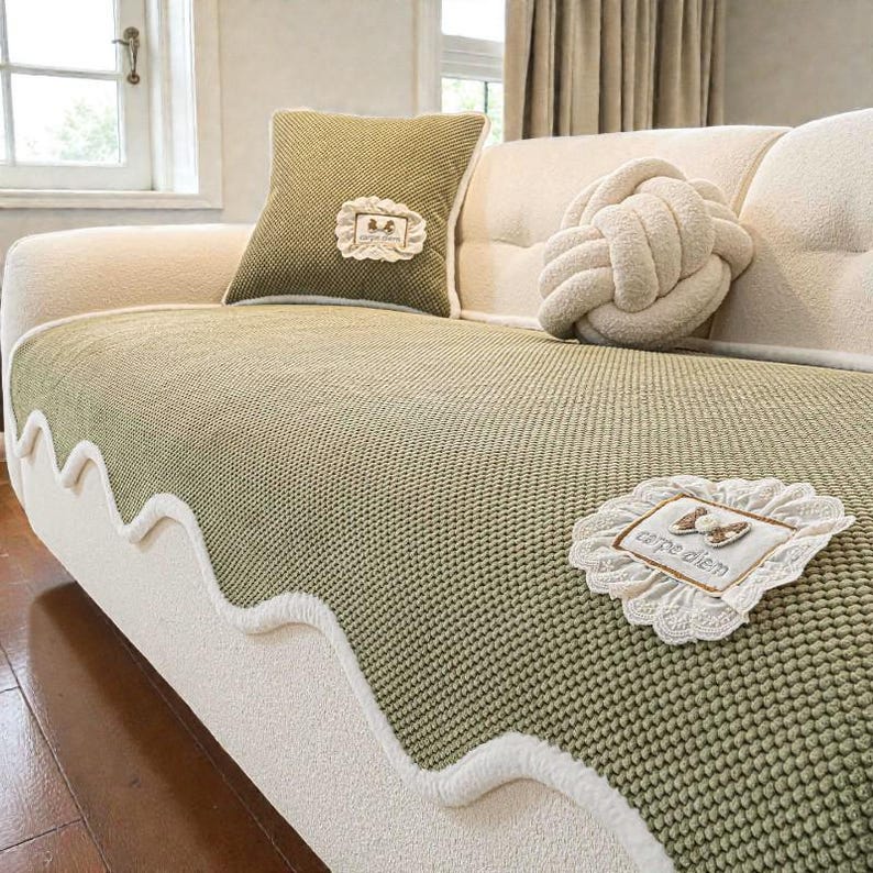 Pu&ograve; includere: Un divano color crema con un copriletto in maglia verde e un cuscino abbinato. Il copriletto ha un bordo bianco ondulato. Sono visibili anche un cuscino decorativo a nodo e una toppa quadrata con la scritta "carpe diem".
