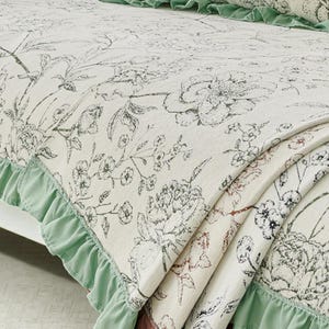 May include: A cream-colored floral patterned bedspread with a sage green ruffled edge. The bedspread features a large-scale floral design in shades of gray and brown, creating a vintage aesthetic. The fabric is folded, showing the texture and layers.