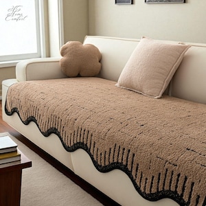 Może przedstawiać: Brązowa narzuta na sofę z czarnym, falistym brzegiem i czarnymi pionowymi liniami. Narzuta jest na kremowej sofie, z dwiema pasującymi poduszkami. Logo "HG Home Comfort" znajduje się w lewym górnym rogu.