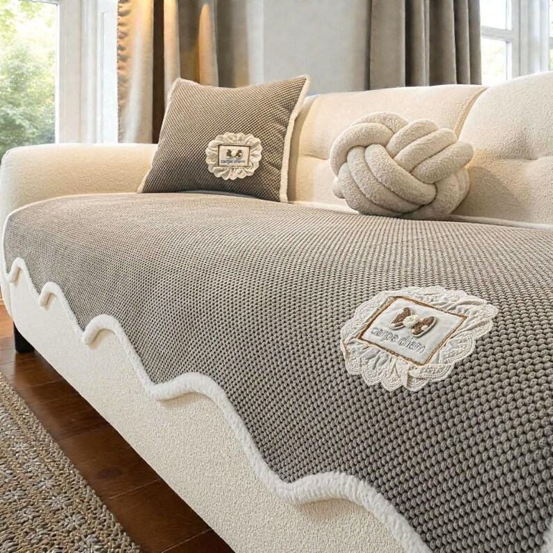 Pu&ograve; includere: Un divano beige con un copriletto in maglia grigia e un cuscino abbinato. Il copriletto ha un bordo bianco smerlato. Un cuscino decorativo a nodo e un cuscino con un design incorniciato sono anche sul divano. Il copriletto dice "carpe diem".