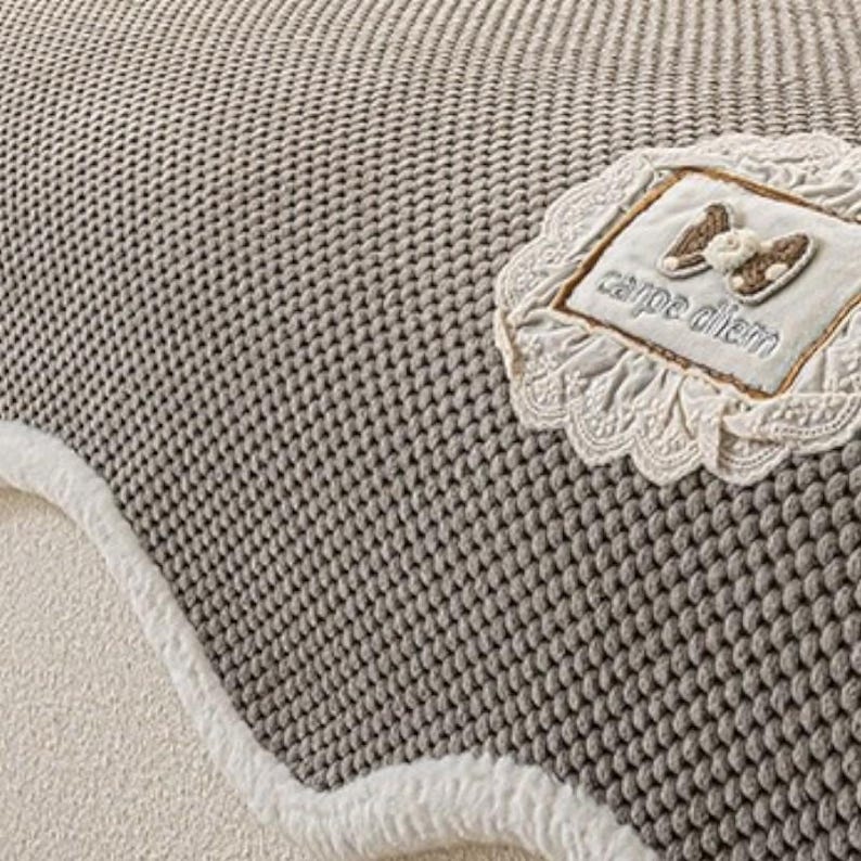 Pu&ograve; includere: Primo piano di una coperta grigia lavorata a maglia con un bordo bianco e soffice. Una toppa decorativa con le parole "carpe diem" e un disegno a fiocco &egrave; cucita sulla coperta. La coperta ha un aspetto strutturato.