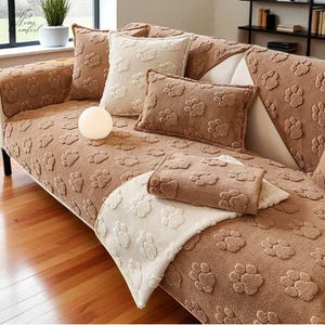 May include: A brown sofa with paw print pattern, featuring matching throw pillows and a blanket. The pillows are in shades of brown and cream. The sofa is in a living room setting with a plant and a lamp.