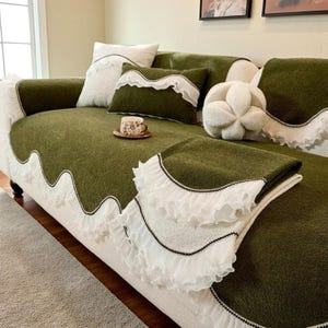 May include: A green sofa cover with white lace trim and matching pillows. The cover has a wavy edge design and is draped over a white sofa. A small teacup and saucer sit on the sofa.