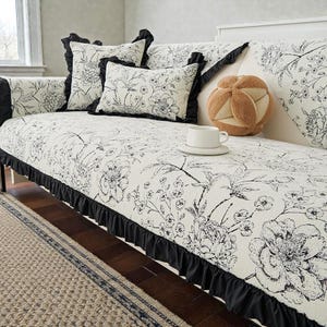 May include: A sofa cover featuring a black and white floral print with a black ruffled trim. The set includes matching square and rectangular throw pillows, and a tan, multi-lobed plush ball pillow sits on the seat next to a white ceramic mug.
