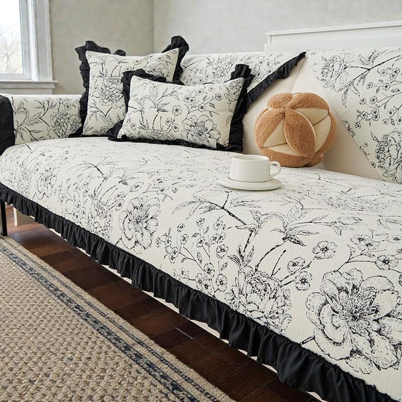 May include: A sofa cover featuring a black and white floral print with a black ruffled trim. The set includes matching square and rectangular throw pillows, and a tan, multi-lobed plush ball pillow sits on the seat next to a white ceramic mug.