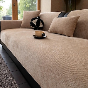 May include: A beige sofa with matching throw pillows and a black and white decorative ball. A beige teacup and saucer sit on the sofa. The sofa has a dark brown base and is placed near a window.