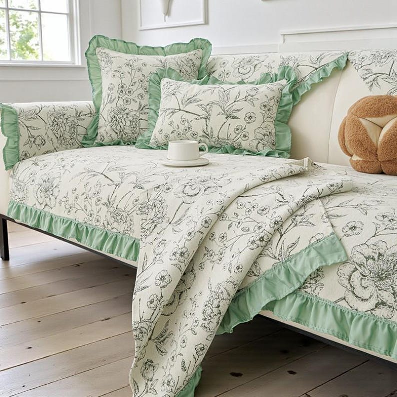 May include: A white sofa with floral patterned cushions and a matching throw. The cushions and throw have a black and white floral design with green ruffled edges. A white teacup and saucer sit on the sofa.