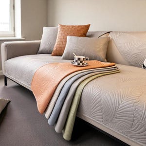May include: A modern grey sofa with decorative cushions and a folded throw blanket. The cushions include a woven brown leather cushion and two grey cushions with a leaf pattern. The throw blanket is folded over the sofa in shades of orange, grey, and green.