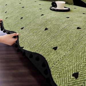 May include: A green sofa cover with a herringbone pattern and black heart accents. The cover has a black pom-pom trim. A white teacup and saucer sit on the cover.