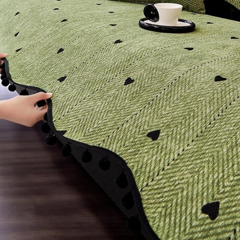 May include: A green sofa cover with a herringbone pattern and black heart accents. The cover has a black pom-pom trim. A white teacup and saucer sit on the cover.