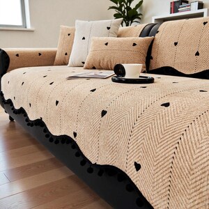 May include: A beige sofa cover with a black heart pattern and a black scalloped edge. The sofa has several matching throw pillows with heart designs. A coffee cup sits on a black saucer on the sofa.