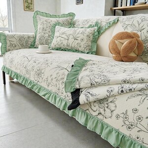 May include: Off-white sofa cover with a floral pattern in black and gray. The cover has a green ruffle trim and matching throw pillows. A decorative brown ball-shaped pillow and a white teacup are also visible.
