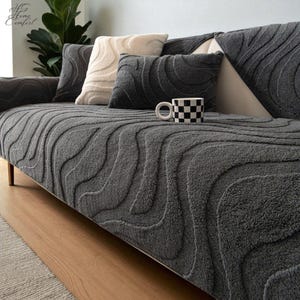 May include: A gray sofa cover with a textured, wave-like pattern. The sofa is adorned with several pillows in shades of gray and cream. A black and white checkered mug sits on the sofa. The Home Comfort logo is visible in the upper left corner.