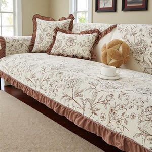 May include: A cream-colored sofa cover featuring a brown floral print and matching brown ruffled trim. The set includes a square throw pillow and a rectangular lumbar pillow, both adorned with the same floral pattern and ruffled edges.