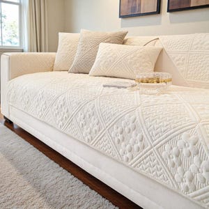 Könnte beinhalten: Ein cremefarbenes Sofa mit einer gesteppten Decke und passenden Wurfkissen. Die Decke hat ein geometrisches Muster mit Blumen- und Diamantformen. Eine kleine dekorative Box und ein Buch befinden sich auf dem Sofa.