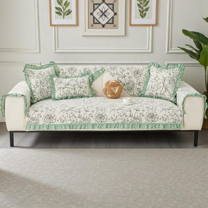 May include: A white sofa with a floral patterned cover and matching throw pillows. The cover and pillows have a black and white floral design with green ruffle trim. Decorative pillows and a small cup and saucer are on the sofa.