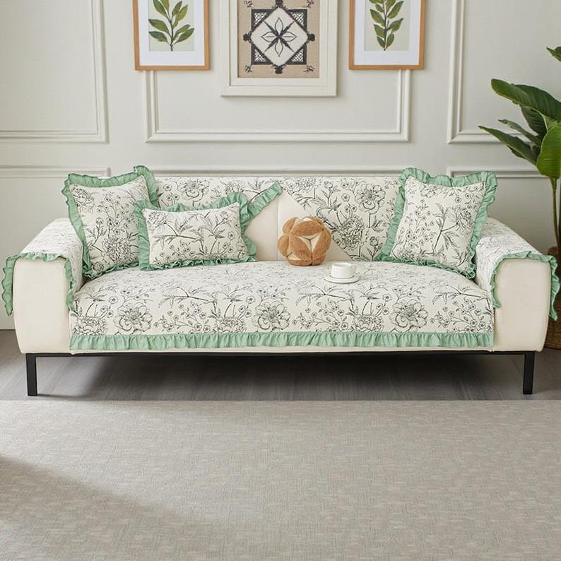 May include: A white sofa with a floral patterned cover and matching throw pillows. The cover and pillows have a black and white floral design with green ruffle trim. Decorative pillows and a small cup and saucer are on the sofa.