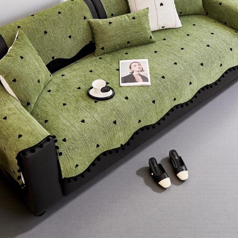 May include: A green sofa with black leather accents and matching throw pillows. The sofa is covered with a green patterned throw blanket with small black hearts. A magazine, a teacup, and a pair of black and white shoes are on the sofa.