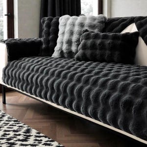 Könnte beinhalten: Ein modernes Sofa mit einem dunkelgrauen, strukturierten Kunstfellbezug und passenden Kissen. Das Sofa hat einen weißen Rahmen und schwarze Beine. Ein schwarz-weiß gemusterter Teppich ist im Vordergrund zu sehen.