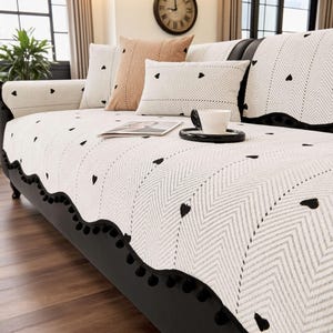 May include: A black sofa with a white and gray patterned cover featuring black heart accents. Decorative pillows in white, pink, and gray complement the sofa. A black trim with black pom-poms lines the bottom edge of the cover.